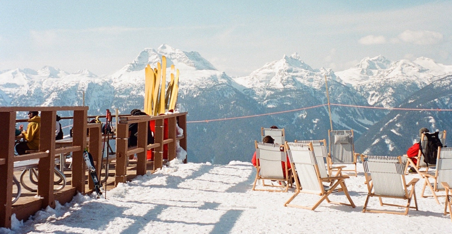 Snowy mountain top with wooden deck, chairs, and people enjoying the view.